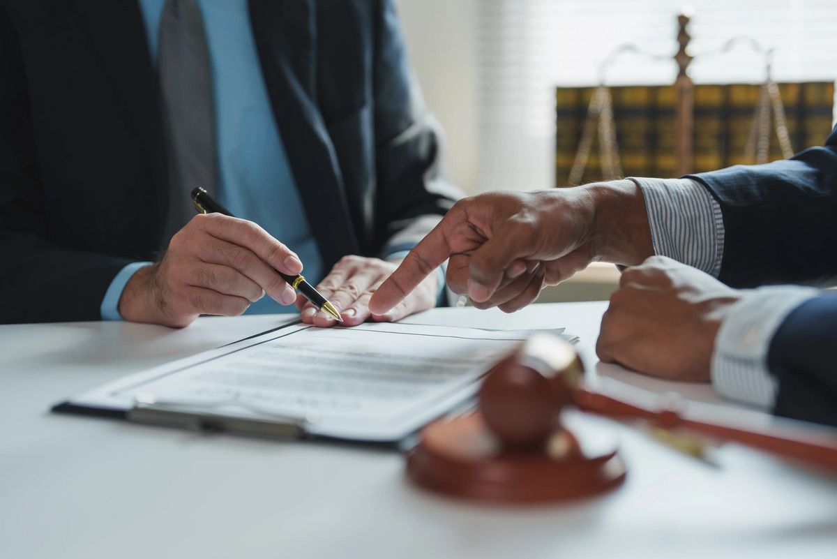 Two lawyers are discussing contract details, pointing at specific clauses with a pen, with a gavel and scales of justice in the background, symbolizing legal proceedings and agreement