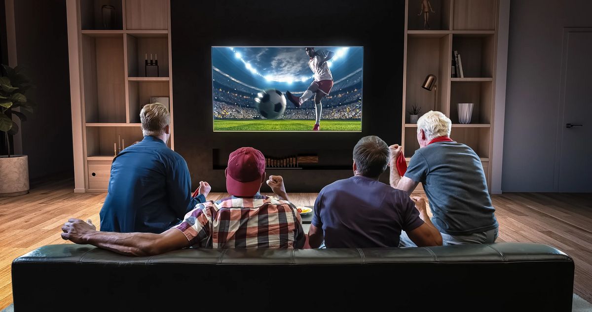 Group of fans are watching a soccer moment on the TV and celebrating a goal, sitting on the couch in the living room.