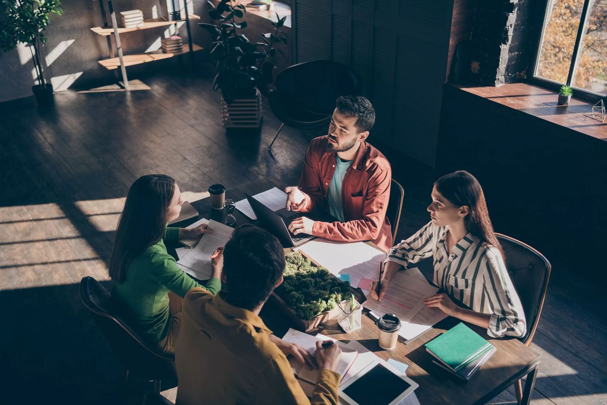 Four busy experienced businesspeople wearing casual formal-wear discussing delegation company growth income at modern industrial loft wooden interior workplace workstation